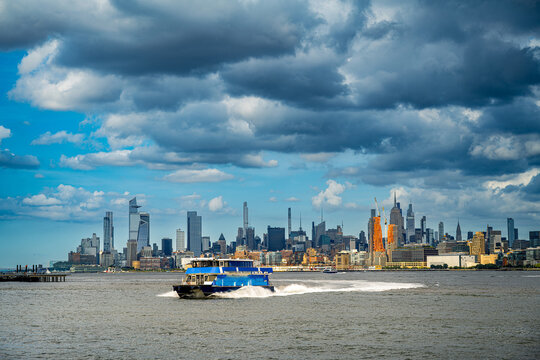 Commuter ferry moves across Hudson River below Manhattan skyline. Blue passenger vessel produces a long wake beneath Midtown towers and layered gray clouds.