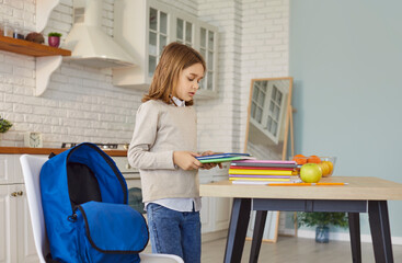 Child arranges a stack of colorful school books beside a blue backpack on the kitchen table, preparing notebooks and supplies for school in a bright home. She packs books for school now. © Studio Romantic