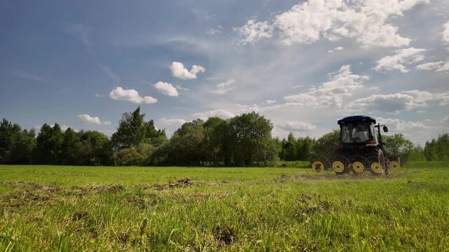 Farm Tractor Tedding Hay with Rotary Rake in Green Field, Blue Sky and Forest, Rural Countryside Agricultural Machinery Working on Summer Farmland