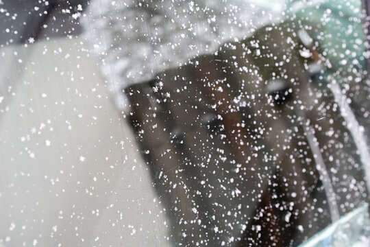 Close-up of a car window covered with delicate snowflakes. The crystalline patterns stand out against the reflective surface, where faint outlines of the surroundings appear blurred.