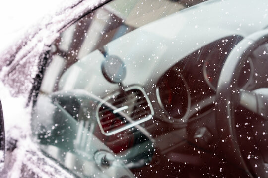 Close-up of a car window covered with delicate snowflakes. The crystalline patterns stand out against the reflective surface, where faint outlines of the surroundings appear blurred.