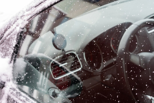 Close-up of a car window covered with delicate snowflakes. The crystalline patterns stand out against the reflective surface, where faint outlines of the surroundings appear blurred.