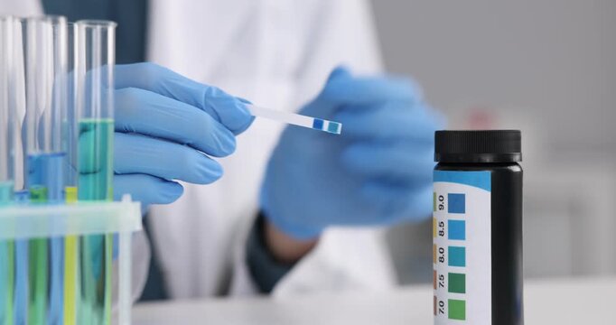 Scientist holding PH test strip with color table at white table in laboratory, closeup