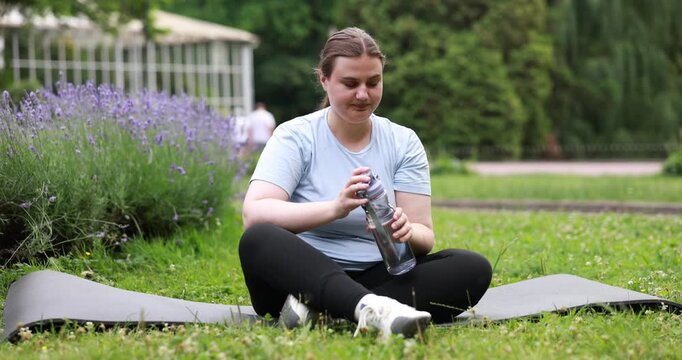 Overweight woman drinking water from bottle and exercising on fitness mat in park