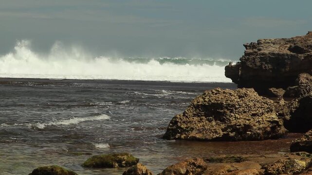 Powerful ocean waves crashing over a coral reef near a rocky coastline under a cloudy sky