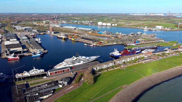 Aerial wide angle panoramic view of a bustling seaport harbor with industrial shipyards luxury mega yachts cargo vessels and a sea dike embankment under a clear blue sky