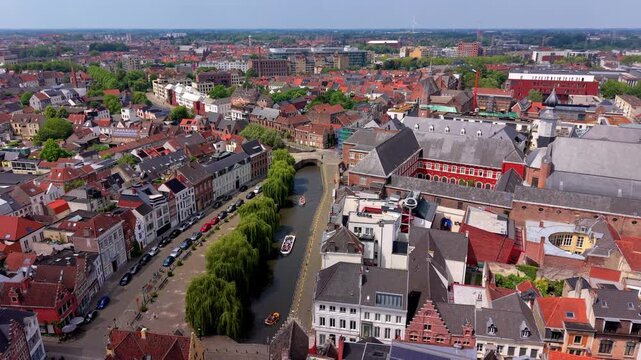 Aerial wide angle panoramic view over a scenic city river canal with tourist tour boats traditional architecture red tiled roofs and a row of green trees under a summer horizon sky