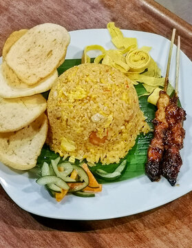 Authentic Indonesian nasi goreng served with grilled chicken satay, prawn crackers, egg ribbons, pickled vegetables and banana leaf on a white plate.
