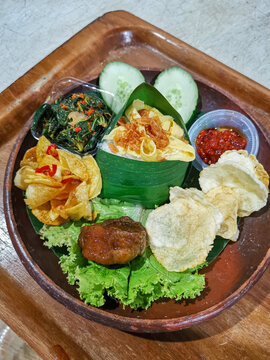 Authentic Indonesian mixed rice served with beef rendang, spicy cassava leaves, sambal, egg strips, crackers and fresh vegetables on a rustic clay plate.