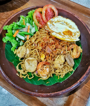 Authentic Indonesian mie goreng seafood served with shrimp, fried egg, fish balls, meatballs, pickled vegetables and banana leaf in a rustic clay plate.