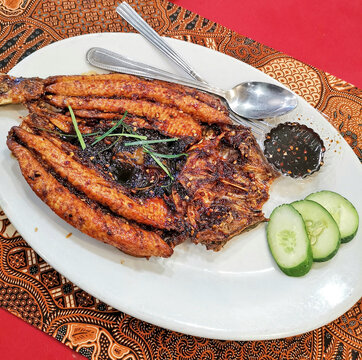Authentic Indonesian Bandeng Kropok featuring crispy butterflied milkfish served with sweet soy chili dipping sauce and cucumber on a white plate over traditional batik tablecloth.