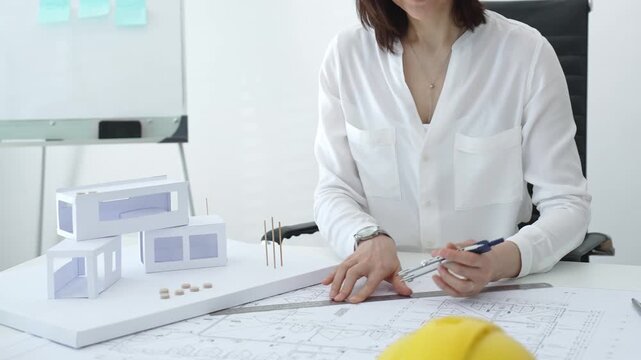 Professional female architect working on blueprints and scale models at a bright office desk, concentrating on urban planning, structural design and project development