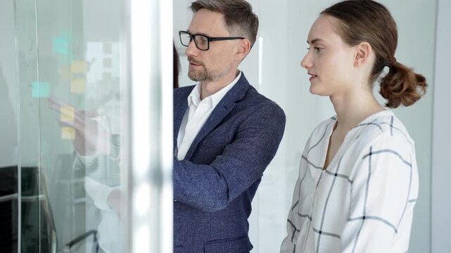 Business team collaborating on a project, analyzing data on a tablet, planning strategy with sticky notes on a glass whiteboard in a modern office meeting