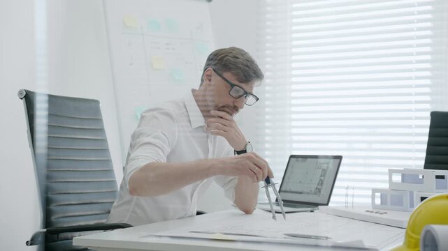 Male architect in modern office creating architectural design blueprint on paper and working on laptop, developing new building project with models on white desk