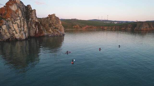 Shamanka Rock, Lake Baikal on a summer evening. Tourists on SUP boards are paddling in the bay.