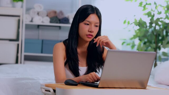 Young asian woman using laptop on bed in bedroom for remote work on computer at home serious focused indoor lifestyle with plant shelf notebook pillow window daylight casual setup