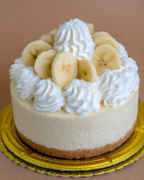 Round banana cheesecake with a graham crust, whipped cream rosettes and banana slices on top, presented on a gold cake board.