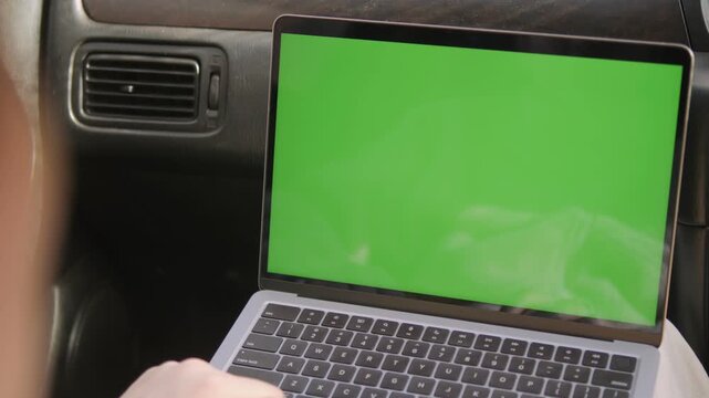 Person works on laptop with green screen inside car. Fingers type on keyboard, screen shows chroma key. Focus on hands and device, background is car interior. Ideal for remote work, travel
