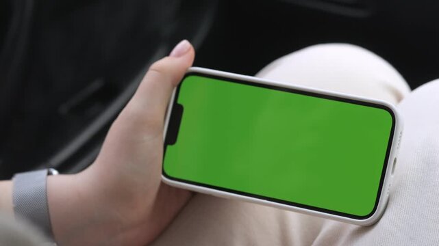 Hand holds phone with green screen inside car. Person wears smartwatch and white pants. Focus on device for app or advertisement use. Ideal for car-related or tech content