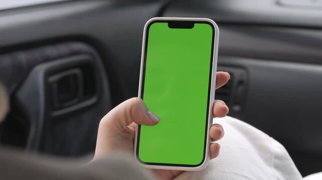 Hand holds smartphone with green screen inside car. Person wears white clothes. Focus on phone screen for overlays. Ideal for mobile app demos or navigation concepts. Realistic car interior setting