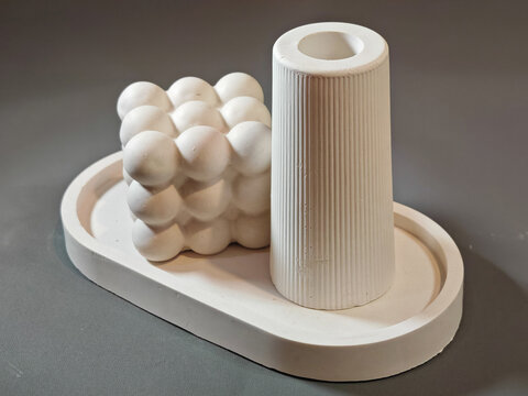 A high-contrast commercial studio shot featuring modern white concrete home accessories including a bubble cube and a fluted cylindrical holder arranged on a matte grey background