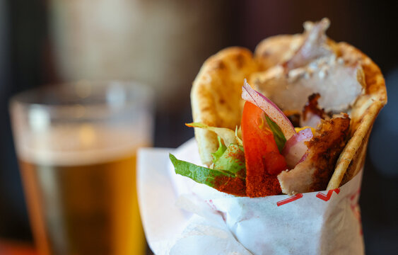 Pita gyro stuffed with tender grilled meat, fresh tomato, crisp lettuce, and creamy tzatziki, served beside a cold frothy beer &mdash; a classic taste of Greek street food and Mediterranean flavor.