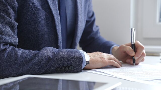 Professional businessman signing contract with silver pen, wearing tailored blue suit, business documents and digital tablet nearby. Business people