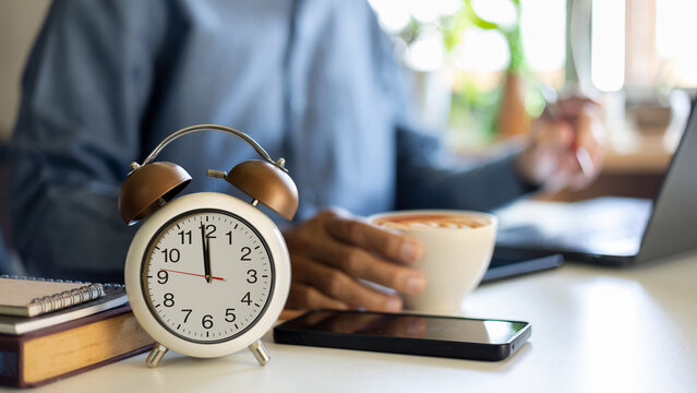 close up alarm clock before 12 PM with asian businessman holding coffee cup using laptop working on desk before taking a break at lunch time