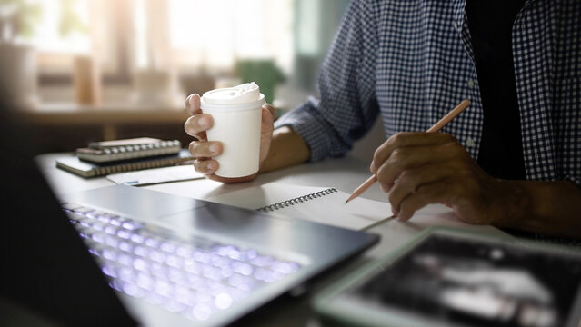 close up shot, asian man holding coffee cup using laptop work on desk at home, taking note, checking e-mail, meeting on line, working at home concept