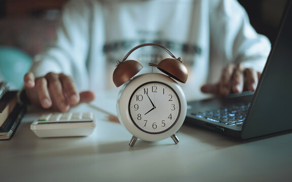 close up shot, focus alarm clock before 8.00 AM. blurred asian man in casual clothes working at home with laptop and calculate expenses from e-commerce business