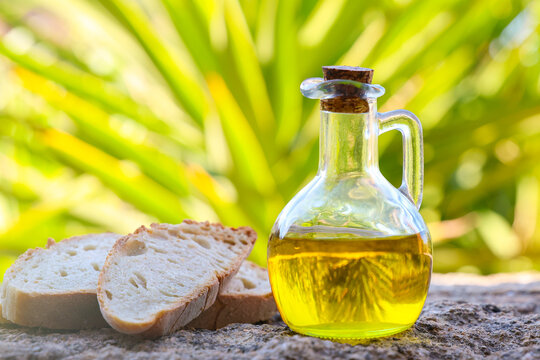 Golden olive oil in a glass bottle with rustic bread slices on a stone surface in Crete, Greece, framed by green yucca leaves and warm Mediterranean sunlight.