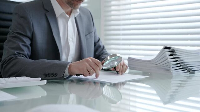 Business professional meticulously inspecting a contract or official document with a magnifying glass and pen while working at a desk with a stack of paperwork