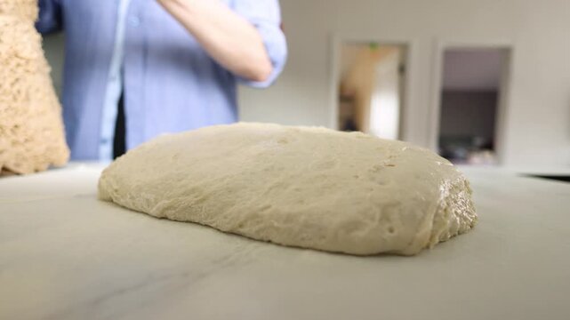 Proofed bread dough rests on floured counter in bright home kitchen as blurred baker shapes and lifts dough nearby, with soft daylight, shallow focus, warm domestic baking mood, with bread, dough
