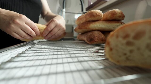 Hands press torn bread onto wire cooling rack beside stacked baked loaves in soft kitchen light, showing soft crumb, golden crust, warm domestic setting, shallow focus, rustic texture, and careful