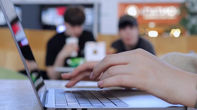 Hand typing on laptop keyboard. Background shows blurred people in cafe setting. Person focused on work, calm and concentrated. Ideal for remote work, freelancing, or online business scenes