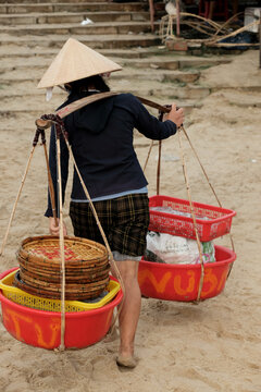 A local woman carrying goods with a traditional shoulder pole while walking on the sandy beach of a fishing village.