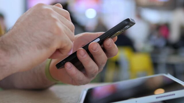 Hand taps smartphone screen in public space. Fingers scroll through phone with focus. Another device lies nearby on table. Person appears calm and engaged with device. Suitable for tech, communication