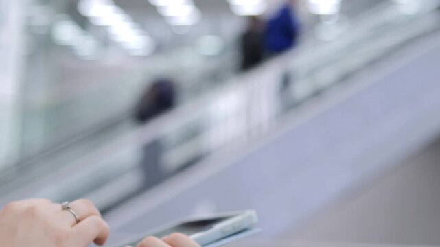 Person holds smartphone while riding escalator. Fingers tap screen, focusing on device. Background shows blurred travelers and modern architecture. Neutral tones, soft lighting, urban travel theme
