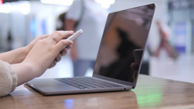Person uses phone while laptop rests on table. Background shows people moving in public space. Reflections visible on laptop screen. Hands interact with both devices simultaneously