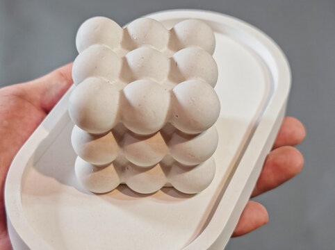 A stock photo showing a modern handcrafted white concrete decorative object with a minimalist geometric bubble structure. The artisan object sits on a matching concrete tray and is held gently