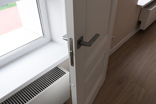 white paneled interior door with dark‑gray handle stands ajar beside white window sill. below it, white radiator with slats is installed, while warm‑toned wooden floor extends into room