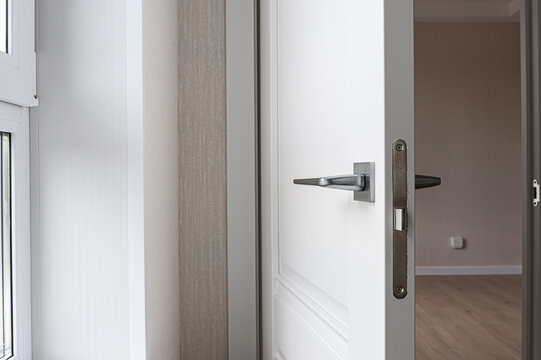 partially open white interior door features sleek silver handle and metallic lock plate. beside it, beige textured wall panel meets doorframe, while window frame is visible to left