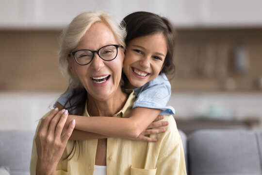 Loving pre-teen 12s granddaughter cuddling from behind, piggyback with love and warmth her older 65s granny, sit on sofa, spend pleasant priceless time together at home. Multigenerational family ties