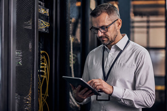 A man in a shirt is looking at a tablet in a server room.