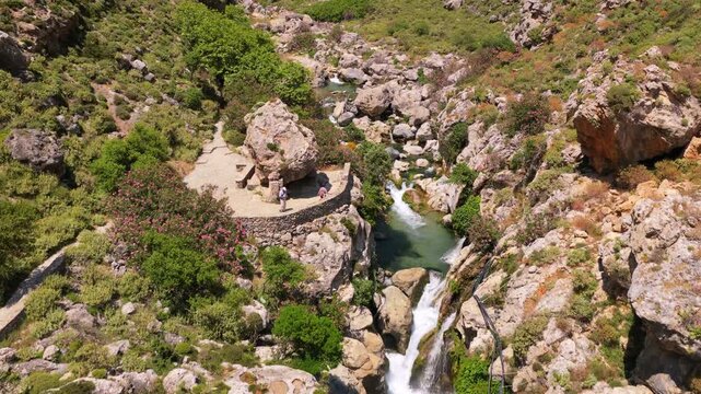 Aerial drone view of a rocky river with small cascades and turquoise pools between boulders and pink oleander, Kourtaliotiko Gorge, Greece
