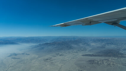 Naklejka premium Flying over the desert. An airplane wing on a blue sky background. Below there are sand dunes, hills, mountains without vegetation, winding riverbeds of dried rivers. Peru. The Nazca Plateau.