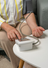 Fototapeta premium Cropped view of woman starting blood pressure measurement with digital monitor while sitting on sofa. Home healthcare and heart health awareness.