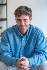 Fototapeta premium Young man in a denim shirt smiles confidently while seated at home, hands clasped in front; casual portrait conveys friendliness, approachability, relaxed confidence and professionalism in a modern 