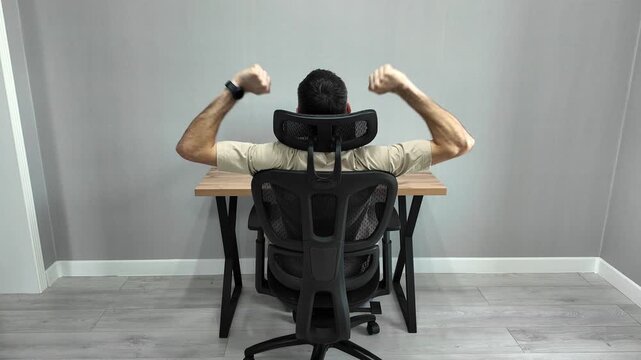 Back view of a young male employee stretching his arms and back while sitting on an ergonomic chair at his office desk during a work break