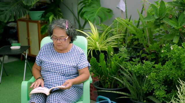 Senior Asian woman relaxing and reading a book in a lush green home garden, elderly retirement lifestyle concept.